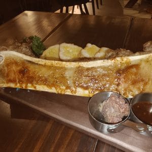 Món xương tủy bò nướng bày trên thớt gỗ ăn kèm bánh mì nướng và sốt