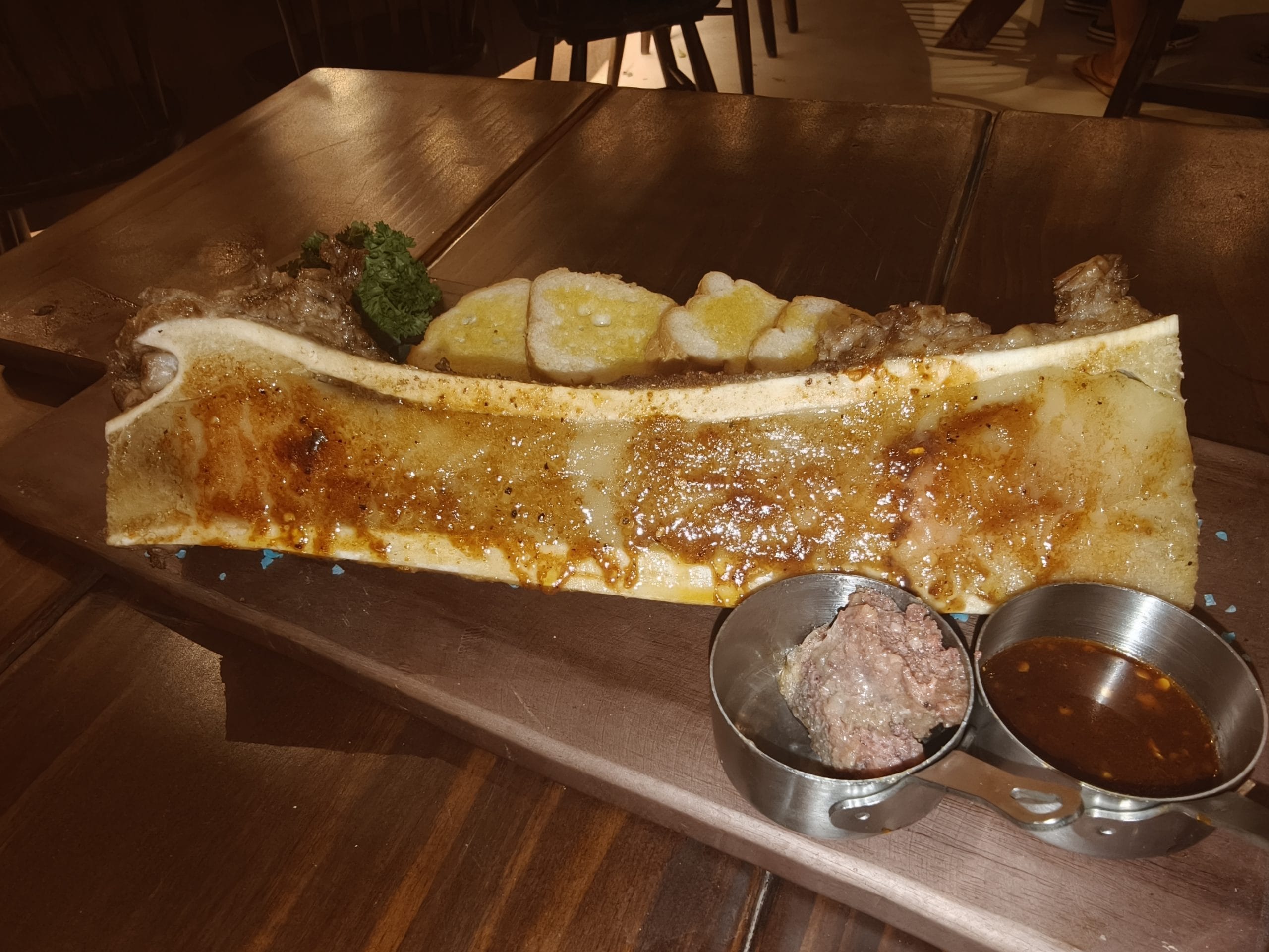 Món xương tủy bò nướng bày trên thớt gỗ ăn kèm bánh mì nướng và sốt